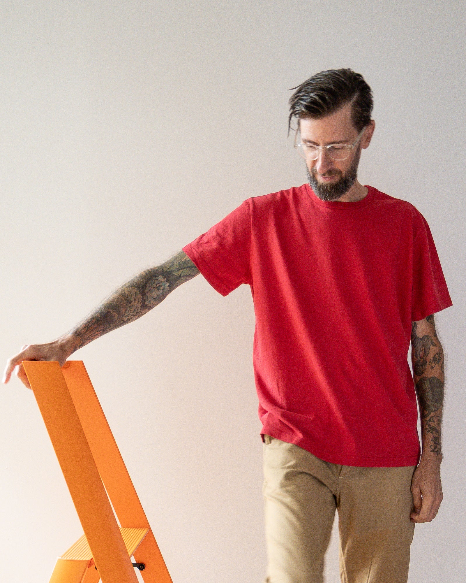 Man wearing a red t-shirt and beige pants standing next to an orange ladder against a plain background