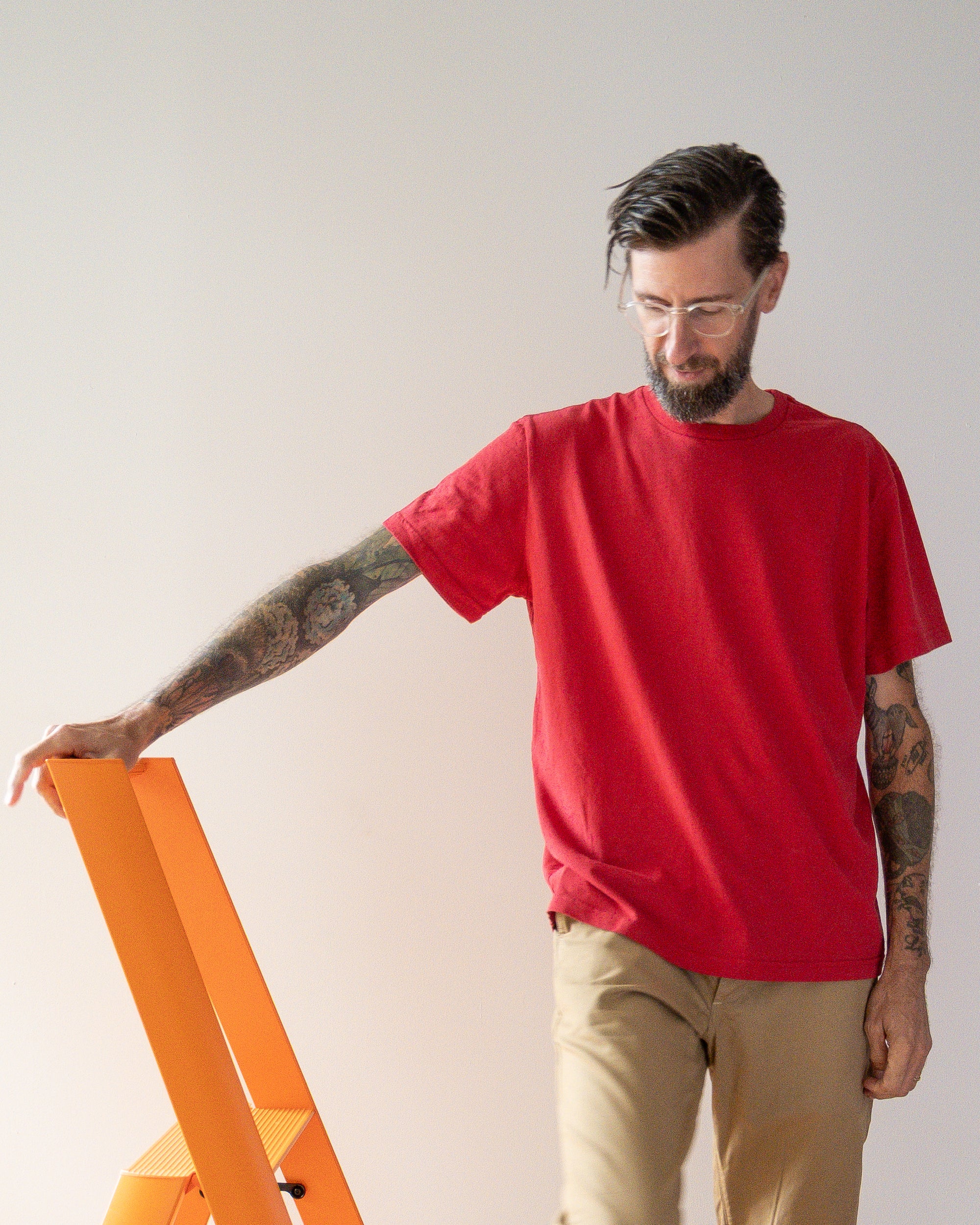 Man wearing a red t-shirt and beige pants standing next to an orange ladder against a plain background